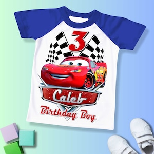 May include: White t-shirt with a blue raglan sleeve design. The shirt features a red race car with the number 3 on the hood, checkered flags, and the text "Caleb Birthday Boy".