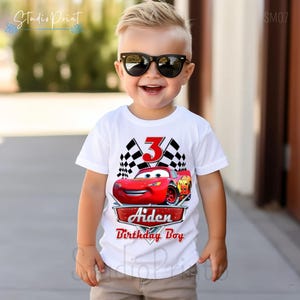 Puede incluir: Camiseta blanca con un gr&aacute;fico de coche rojo de Rayo McQueen, banderas a cuadros y el n&uacute;mero 3. El texto "Aiden Birthday Boy" est&aacute; impreso debajo del coche. Un ni&ntilde;o peque&ntilde;o lleva la camiseta y sonr&iacute;e.