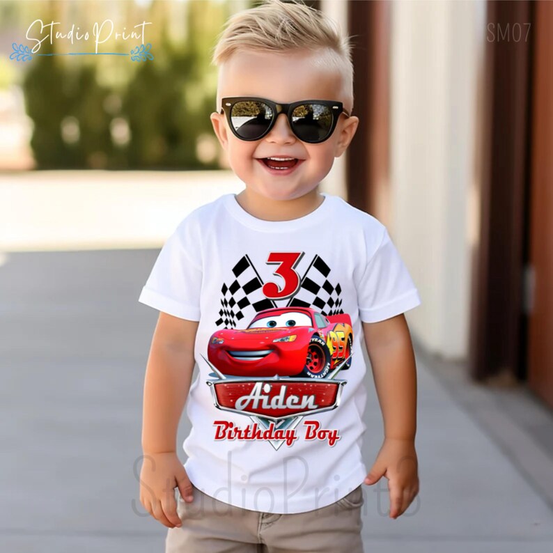 Puede incluir: Camiseta blanca con un gr&aacute;fico de coche rojo de Rayo McQueen, banderas a cuadros y el n&uacute;mero 3. El texto "Aiden Birthday Boy" est&aacute; impreso debajo del coche. Un ni&ntilde;o peque&ntilde;o lleva la camiseta y sonr&iacute;e.