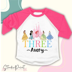 May include: White and pink raglan-style t-shirt featuring Disney princesses and the word "THREE" in colourful letters. The name "Avery" is below the word "THREE". The shirt has three-quarter sleeves and a round neckline. The Studio Print logo is at the bottom.
