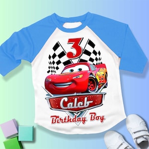 May include: A white and blue baseball t-shirt with a red race car and checkered flags. The number 3 is in red and the text "Caleb Birthday Boy" is in silver.
