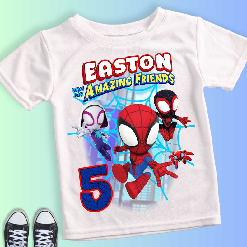以下が含まれることがあります： 「EASTON and his AMAZING FRIENDS」の文字と、スパイダーマンなどのキャラクターの漫画の画像がプリントされた白いTシャツ。数字の「5」も表示されています。Tシャツは半袖です。