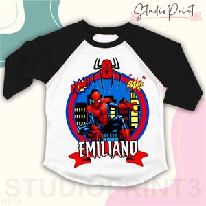 May include: Black and white baseball tee shirt with red and blue Spiderman graphic. The graphic includes a cityscape, a spider, and the words "POW!" and "BAM!" The shirt also has the name "EMILIANO" in red letters.