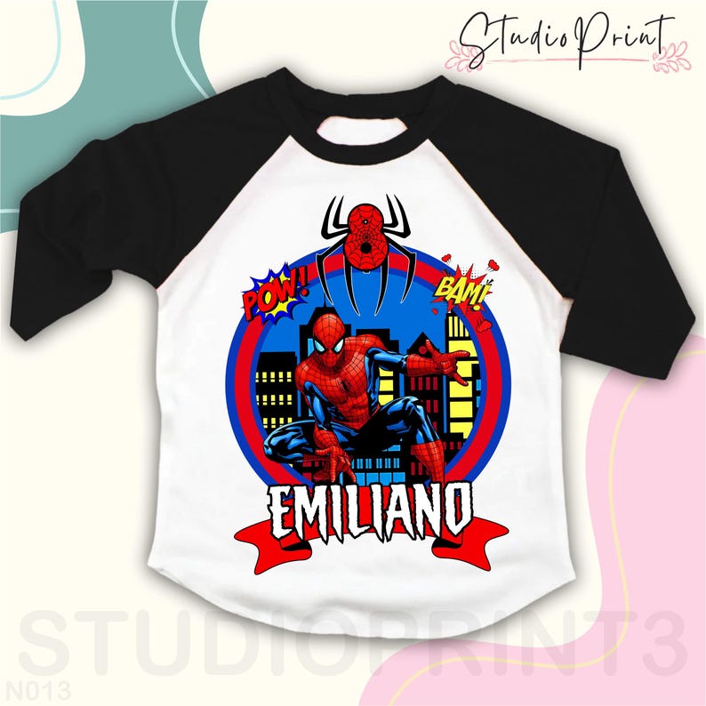 May include: Black and white baseball tee shirt with red and blue Spiderman graphic. The graphic includes a cityscape, a spider, and the words "POW!" and "BAM!" The shirt also has the name "EMILIANO" in red letters.