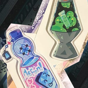 Peut inclure: Deux breloques en acrylique. L'une est une bouteille bleue avec le texte "MeFan!" et du texte japonais, et un visage de dessin animé. L'autre est un design de lampe à lave avec des blobs verts et un tube à essai. Les deux ont une attache porte-clés.