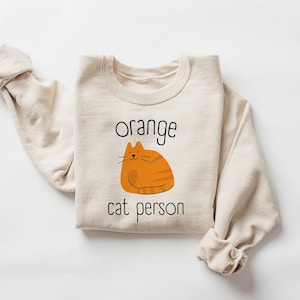 May include: A beige sweatshirt with the text "orange cat person" and a cartoon orange cat illustration.