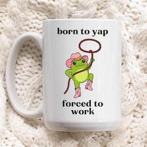 May include: White ceramic mug featuring a green frog in a pink cowboy hat and boots, holding a lasso. The mug is printed with the words "born to yap" and "forced to work". The mug has a large handle.