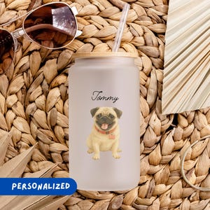 May include: A frosted glass tumbler with a wooden lid and straw, featuring a pug illustration and the name "Tommy." The tumbler has a personalized design. Sunglasses and a gold bracelet are also visible.