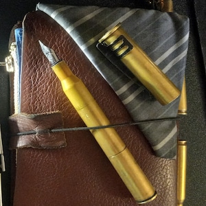 Puede incluir: Un diario de cuero marrón con una tela rayada en negro y gris metida dentro. Un bolígrafo dorado y dos casquillos de bala dorados también están dentro del diario.