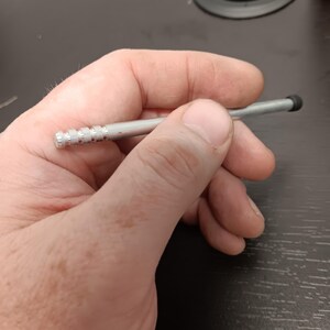 May include: A silver stylus with a black rubber tip. The stylus has a ridged design near the tip and is held in a person's hand. The stylus is designed for use with a touchscreen device.