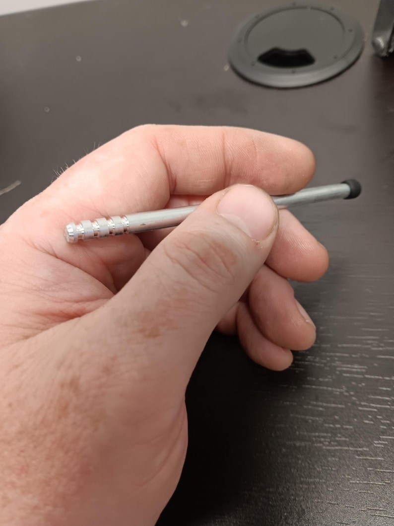 May include: A silver stylus with a black rubber tip. The stylus has a ridged design near the tip and is held in a person's hand. The stylus is designed for use with a touchscreen device.