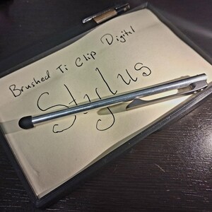 May include: A brushed Ti clip digital stylus rests on a notepad with the word "Stylus" handwritten on it. The notepad is inside a black frame, and a pen is visible in the background. The stylus is silver with a black tip.