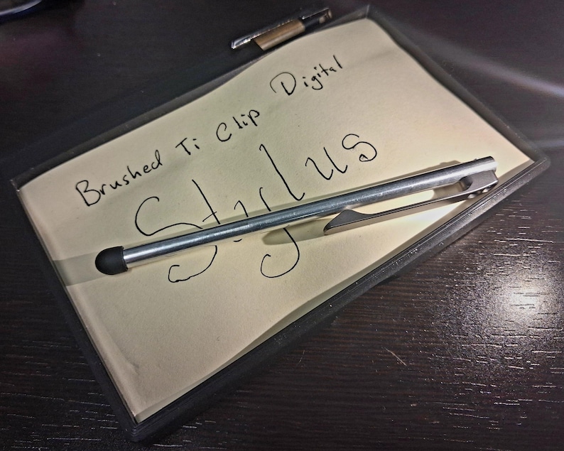 May include: A brushed Ti clip digital stylus rests on a notepad with the word "Stylus" handwritten on it. The notepad is inside a black frame, and a pen is visible in the background. The stylus is silver with a black tip.