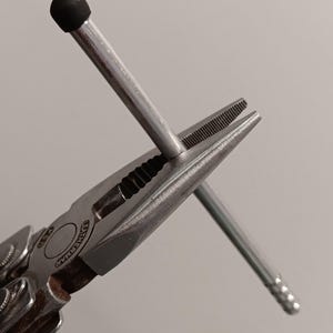 May include: A multi-tool pliers with a silver finish, holding a matchstick with a black tip. The pliers are open, gripping the matchstick. The Leatherman logo is visible on the tool.