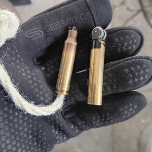 May include: A brass bullet casing with a white wick attached, next to a small, brass-colored lighter. The items are held in a black glove with a textured grip.