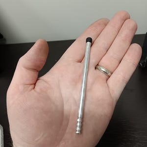May include: A silver stylus with a black tip, held in a hand. The stylus is approximately 5 inches long and has a ridged design near the base. The background is a neutral color.