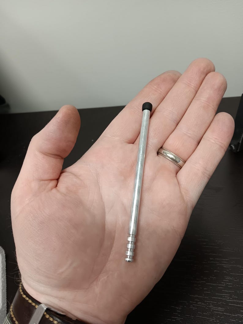 May include: A silver stylus with a black tip, held in a hand. The stylus is approximately 5 inches long and has a ridged design near the base. The background is a neutral color.