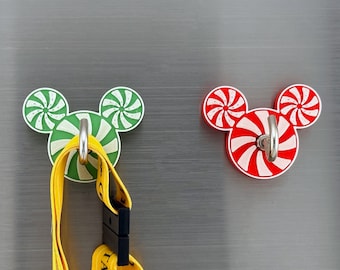 May include: Two decorative hooks shaped like Mickey Mouse ears. The hooks are made of white plastic with a red and white peppermint swirl design and a green and white peppermint swirl design. The hooks are attached to a silver surface.