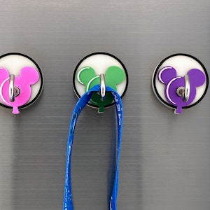 May include: Three round, white, magnetic hooks with a Mickey Mouse head design. The hooks are pink, green, and purple. A blue lanyard is hanging from the green hook.