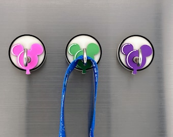 May include: Three round, white, magnetic hooks with a Mickey Mouse head design. The hooks are pink, green, and purple. A blue lanyard is hanging from the green hook.