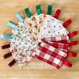 Pode incluir: Um conjunto de dez etiquetas de presente de tecido com padrões diferentes. As etiquetas são amarradas com fita de várias cores, incluindo vermelho, verde e azul. Os padrões incluem estampas florais, xadrez e bolinhas.