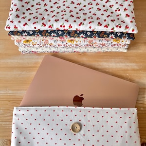 Peut inclure: Un ordinateur portable dans une housse pour ordinateur portable en tissu blanc avec des cœurs rouges. La housse a une fermeture à bouton. L'ordinateur portable est de couleur rose gold et a un logo Apple blanc.