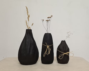 Könnte beinhalten: Drei schwarze Holzvasen in verschiedenen Größen, jede mit getrockneten Pflanzen. Die Vasen haben eine tropfenförmige Form und sind von groß nach klein angeordnet. Die mittlere Vase ist mit Bindfaden gebunden.