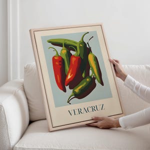 May include: A framed art print with a vibrant illustration of chili peppers in red, green, and yellow hues, set against a pale blue backdrop. The word "VERACRUZ" is printed in teal at the bottom. The frame is a light wood colour.