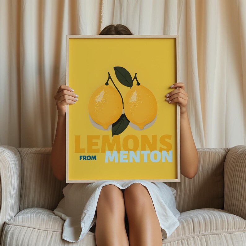A woman is sitting on a couch, holding up a framed poster with a yellow background and an illustration of two lemons.