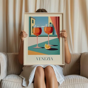A person is sitting on a couch holding a framed picture of two wine glasses with orange slices in front of them, with the word "VENEZIA" written below the image.