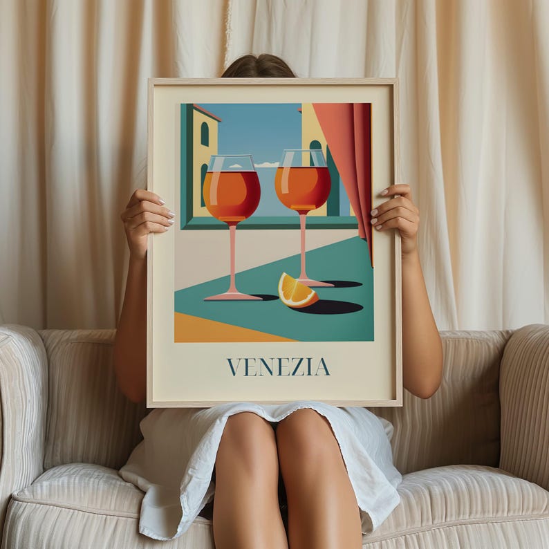 A person is sitting on a couch holding a framed picture of two wine glasses with orange slices in front of them, with the word "VENEZIA" written below the image.