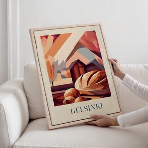 May include: A framed art print with a geometric illustration of a landscape, bread, and the word "HELSINKI" in teal. The artwork uses warm tones of orange, brown, and beige, with a light wooden frame.