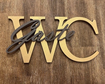Könnte beinhalten: WC-Schild aus Holz mit dem Wort "Gäste" in dunkelbrauner Schrift über den goldfarbenen Buchstaben. Das Schild befindet sich auf einer Holzoberfläche. Ein dekoratives Stück für ein Badezimmer oder Gästezimmer.