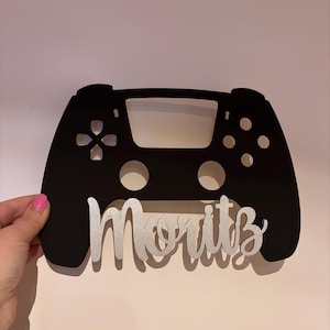 Peut inclure: Silhouette noire d'une manette de jeu vidéo avec le nom "Moritz" écrit en paillettes argentées sur le devant.