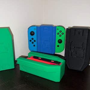 May include: Three 3D printed cases for a Nintendo Switch console. The cases are green, black, and blue and green. The cases have a design on the front that includes the text "BACK2BACK" and "LIFT".
