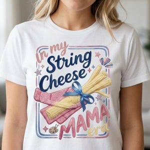 Könnte beinhalten: Weißes T-Shirt mit einem Grafikdesign mit dem Text "In my String Cheese Mama Era". Das Design zeigt Illustrationen von Käsefäden, einem rosa Schal und einer blauen Schleife.