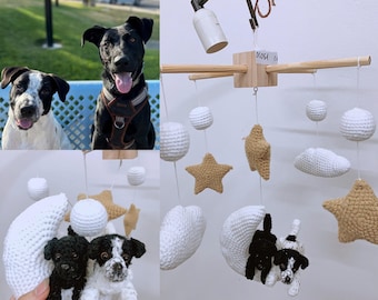 May include: A white crocheted mobile with two crocheted dogs, two stars, two clouds, and two white balls hanging from a wooden frame. The mobile is hanging from a black metal hook. The text "DOGSA" is written on the wooden frame.