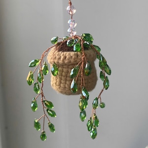 May include: A hanging decorative plant featuring a small, woven tan pot filled with brown material. Green crystal leaves cascade from the pot, attached to copper-colored wire. Clear and pink beads are strung above the pot, creating a unique, handcrafted ornament.