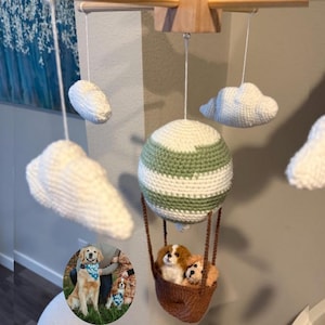 May include: A handmade baby mobile featuring a crocheted hot air balloon with a green and white striped design, a brown basket, and two small dogs. The mobile also includes white crocheted clouds.