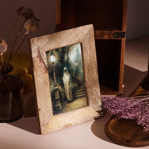 Op de afbeelding: Een houten fotolijst met een donkere, vintage-achtige print van een figuur in een witte jurk die in een mistige straat met lantaarns staat. De lijst heeft een lichtbruine houtkleur.