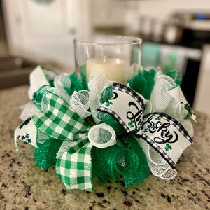 May include: A green and white checkered ribbon, a white ribbon with black and white checkered trim, and a white ribbon with the text "Lucky" in black with green shamrocks are arranged around a clear glass candle holder with a white candle inside. The arrangement is on a green and white speckled surface.