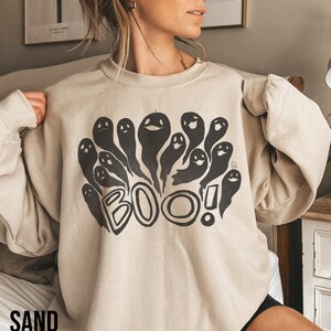 May include: Sand-colored sweatshirt with a black graphic of cartoon ghosts and the word "BOO!" in a playful font. The sweatshirt has long sleeves and a crew neck. The word "SAND" is printed at the bottom of the image.