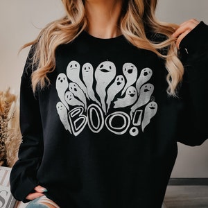 May include: Black sweatshirt with a white graphic of cartoon ghosts and the word "BOO!" in a playful font. The sweatshirt is a crew neck style, perfect for Halloween or spooky season.