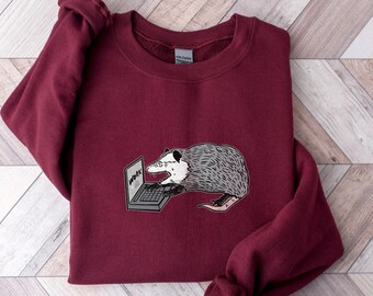 Könnte beinhalten: Burgunderfarbenes Sweatshirt mit einem Cartoon-Opossum-Design. Das grau-weiße Opossum arbeitet an einem Laptop, auf dessen Bildschirm das Wort "WORK" steht. Das Sweatshirt hat eine einfarbige burgunderrote Farbe.
