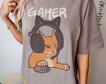 Puede incluir: Una camiseta gris con la palabra "GAMER" impresa en blanco. Debajo de la palabra está impreso un gato naranja de dibujos animados con auriculares y sosteniendo un ratón de ordenador.