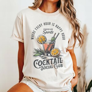 May include: A beige t-shirt with a graphic of a cocktail glass with orange slices and the text "Where every hour is happy hour, Premium Spirits, Cocktail Social Club".