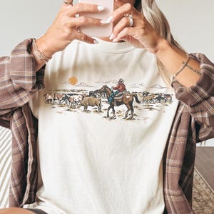 May include: Ivory-colored t-shirt featuring a Western-themed graphic with a cowboy on horseback and cattle. The shirt is paired with a brown plaid shirt, black shorts, and black socks with white stripes. A brown baseball cap is worn.