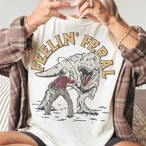 May include: Ivory-colored t-shirt with the text "FEELIN' FERAL" above a graphic of a cowboy wrestling a dinosaur. The person in the image is wearing a brown baseball cap and a brown plaid shirt.