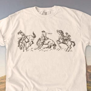 Puede incluir: Camiseta de color marfil con un gr&aacute;fico en tonos sepia de vaqueros montando caballos salvajes. El dise&ntilde;o muestra a tres vaqueros en acci&oacute;n, uno de ellos siendo derribado. La camiseta tiene mangas cortas y cuello redondo. El texto "IVORY" est&aacute; abajo.