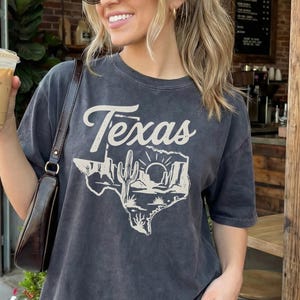 May include: Dark gray t-shirt with the word "Texas" and a desert landscape graphic. The shirt is worn with leopard print pants and a brown purse. The person is holding a coffee drink.